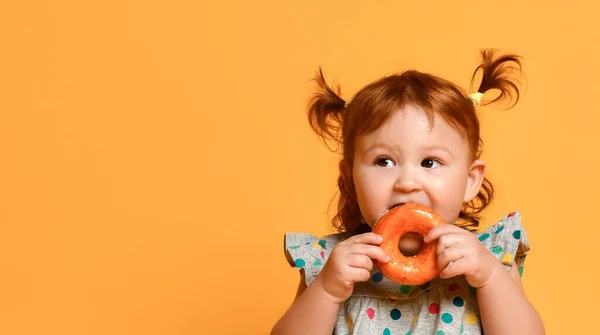 Peuter meisje met twee paardenstaarten, in kleurrijke jurk en witte sandalen. Zittend op de vloer, donut etend, poserend op oranje achtergrond — Stockfoto