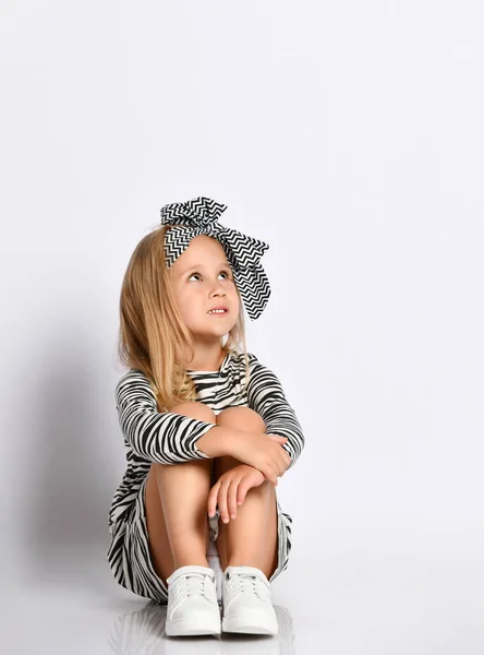 Studio girato bambina in un abito a righe e fascia per capelli. Si siede sul pavimento e guarda verso il luogo per la vostra pubblicità — Foto Stock