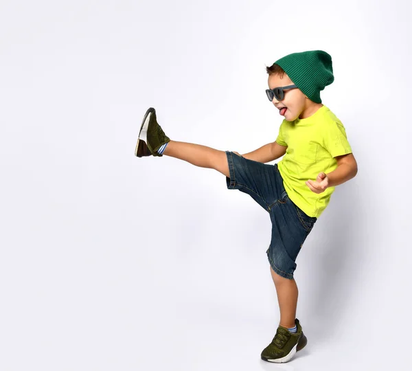 Mały chłopiec w okularach przeciwsłonecznych, zielony kapelusz, żółta koszulka, jeansowe szorty, trampki khaki. Podniesiona noga z języczkiem. Odizolowane na biało — Zdjęcie stockowe