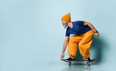 Tişörtlü, turuncu pantolonlu, şapkalı, spor ayakkabılı yaşlı bir adam. Siyah kaykay sürüyor, mavi arka planda yan poz veriyor. Tam uzunluk