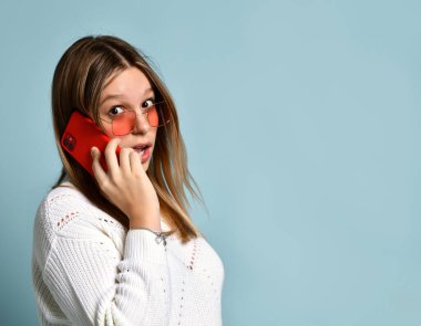 Turuncu güneş gözlüklü ergen, saat, bilezik ve kazak. Telefonda konuşuyor, şaşırmış, mavi arka planda poz veriyor. Kapat.