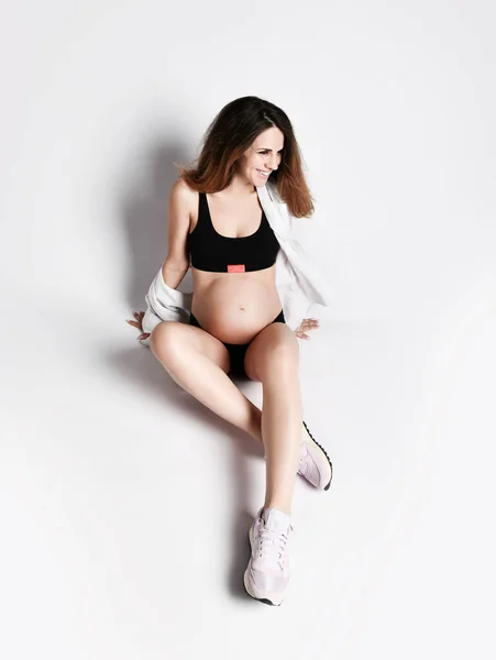 검은 색 스포츠 바지와 윗부분에 임신 한 여성 셔츠, 핑크 운동화. 그는 하얀 바닥에 고립된 채웃고 있었다. 복사 공간 — 스톡 사진