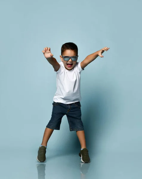 Маленька дитина в сонцезахисних окулярах, біла футболка, джинсові шорти та кросівки хакі. Налякавши тебе, підняв руки, позує на синьому фоні — стокове фото