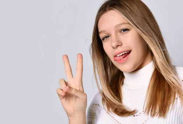 Chica adolescente rubia en suéter y pulsera. Muestra su lengua y su mano victoriosa, posando aislada sobre blanco. De cerca. — Foto de Stock