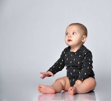 Çıplak ayaklı, siyah giysili küçük bebek. Beyaz stüdyo arka planında izole bir şekilde yerde otururken gülümsüyor. Kapat.
