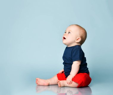 Ginger tişörtlü, kırmızı şortlu, yalınayak. O ağlıyor, mavi arka planda yerde oturuyor. Kapat.