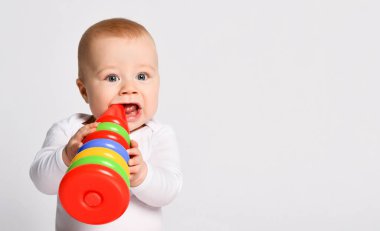 Çıplak ayaklı, takım elbiseli küçük bir çocuk. Çok renkli yığın halkaları oyuncakları çiğniyor, beyaz zeminde izole bir şekilde oturuyor. Kapat.