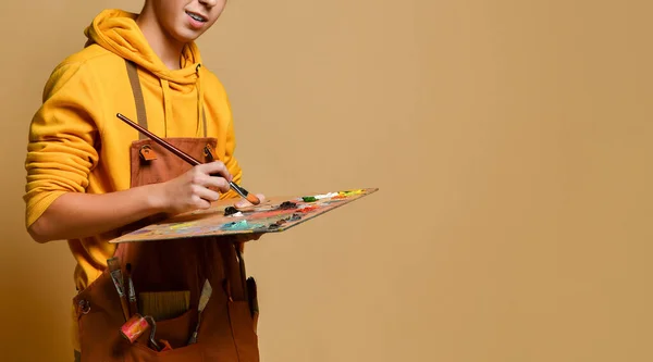 Joven adolescente pintor con capucha y mono amarillo de pie y pintando sobre caballete sobre fondo amarillo — Foto de Stock
