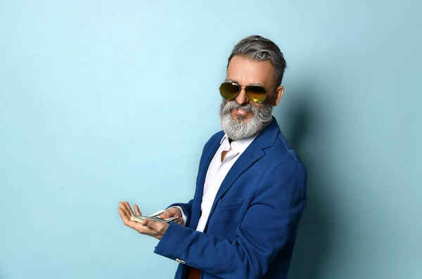 Hombre mayor con camisa blanca, chaqueta, pantalones marrones y gafas de sol. Él mostrando fan de billetes de cien dólares, posando sobre fondo azul — Foto de Stock