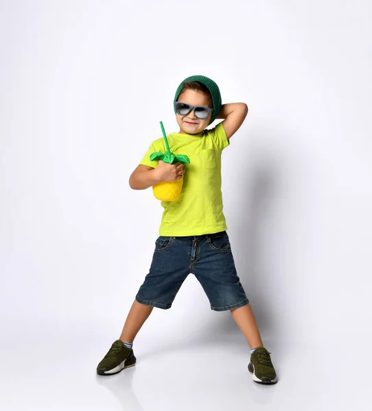 Pojke i solglasögon, grön hatt, gul t-shirt, jeansshorts, khakiskor. Dricker cocktail genom tubuli. Isolerad på vitt — Stockfoto