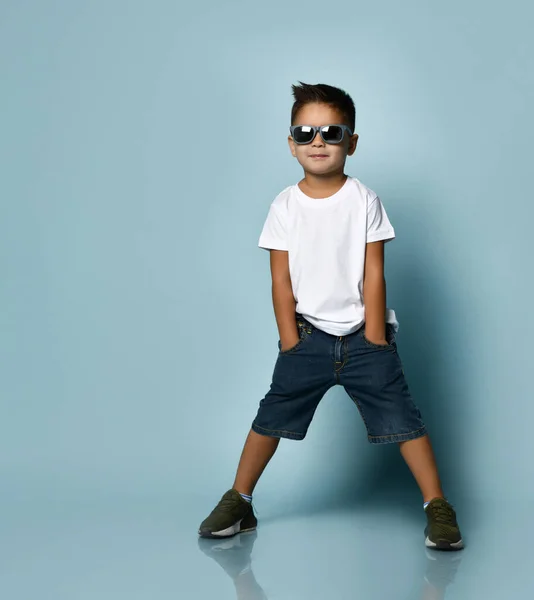 Chłopiec w okularach przeciwsłonecznych, białej koszulce, jeansowych spodenkach i trampkach khaki. Uśmiechnij się, włóż ręce do kieszeni, pozując na niebieskim tle — Zdjęcie stockowe