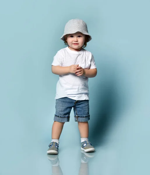 Ginger copil băiat sau fată în tricou alb, pălărie gri, șosete și pantofi, pantaloni scurți denim. Copilul zâmbește, pozând pe fundal albastru — Fotografie, imagine de stoc