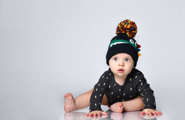 Siyah elbiseli, şapkalı, timsah izli, yalınayak bir çocuk. Gülümsüyor, beyaz zeminde tek başına oturuyor. Kapat.