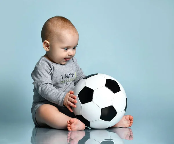 Niño jengibre en traje gris, descalzo. Sostenía la pelota de fútbol, sentado en el suelo sobre fondo azul. Cerrar, copiar espacio —  Fotos de Stock