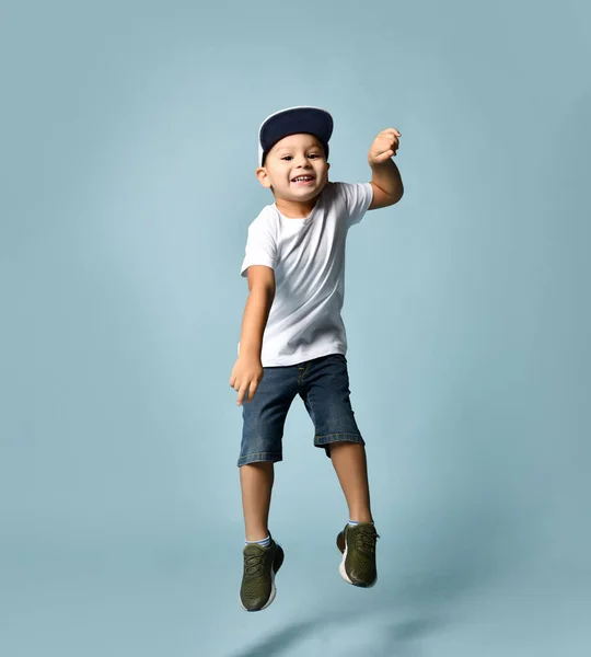 Beyaz tişörtlü, kot şortlu ve haki ayakkabılı küçük esmer çocuk. Zıplıyor, gri başlık takıyor, mavi arka planda poz veriyor