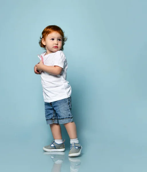 Імбирний малюк хлопчик або дівчинка в білій футболці, шкарпетках та взутті, джинсові шорти. Тримає сонцезахисні окуляри, позуючи збоку на синьому фоні — стокове фото