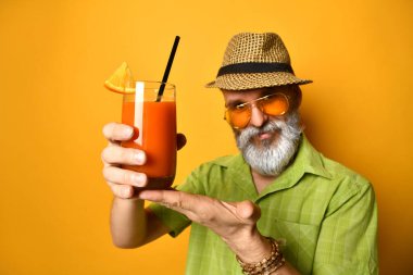 Şapkalı, yeşil gömlekli, güneş gözlüklü yaşlı bir erkek. Gülümseyen ve bir bardak taze meyve suyu sunan, turuncu arka planda poz veren. Kapat.