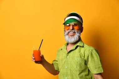 Yeşil güneşlik, gömlek ve güneş gözlüklü sakallı adam. Bir bardak sıkılmış meyve suyu, sıkılmış yumruk, turuncu arka planda poz.