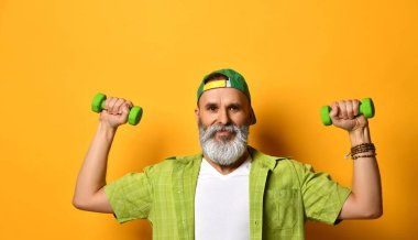 Yeşil şapkalı, tişörtlü, beyaz tişörtlü, bilezik takan yaşlı bir erkek. Gülümsüyor, halterlerle egzersiz yapıyor, turuncu arka planda poz veriyor.