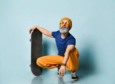 Tişörtlü, güneş gözlüklü, turuncu pantolonlu, şapkalı, spor ayakkabılı yaşlı bir adam. Sana bakıyorum, elinde kaykayla, mavi arka planda oturuyorsun.