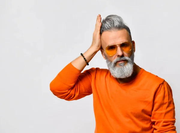 Γέρος άντρας με πορτοκαλί πουλόβερ και γυαλιά ηλίου, βραχιόλι. Φτιάχνει το χτένισμά του, ποζάρει απομονωμένος σε λευκό φόντο. Κλείσε. — Φωτογραφία Αρχείου