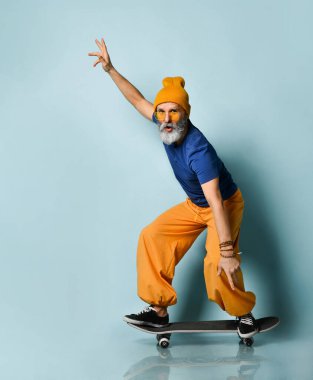 Tişörtlü, güneş gözlüklü, turuncu pantolonlu, şapkalı, spor ayakkabılı yaşlı bir adam. Siyah kaykay sürmek, yumruklarını göstermek, mavi arka planda poz vermek.