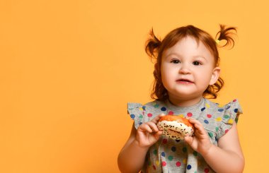 İki at kuyruklu, renkli elbiseli ve beyaz sandaletli küçük kız. Yerde oturmak, donut yemek, turuncu arka planda poz vermek.