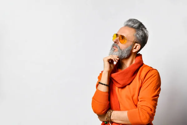 Hombre rico en suéter naranja y bufanda, gafas de sol, pulseras. Se está tocando la barba, posando de lado aislado sobre blanco — Foto de Stock