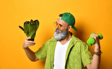 Yeşil şapkalı, gömlekli, beyaz tişörtlü, güneş gözlüklü yaşlı bir adam. Turuncu arka planda iki dambıl ve marul duruyor.