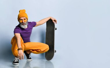 Tişörtlü, güneş gözlüklü, turuncu pantolonlu, şapkalı, spor ayakkabılı yaşlı bir adam. Sana bakıyorum, elinde kaykayla, mavi arka planda oturuyorsun.