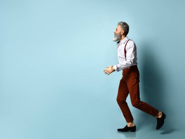 Beyaz gömlekli, kahverengi pantolonlu, pantolon askılı, siyah makosen ayakkabılı yaşlı bir adam. Geriye doğru koşuyor, elini kaldırıyor, mavi arka planda.