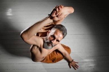 Çıplak gövdeli, kahverengi pantolonlu ve bilezikli yaşlı bir yogi adam esnekliğini gösteriyor. Beyaz ahşap bir zeminde yatarken