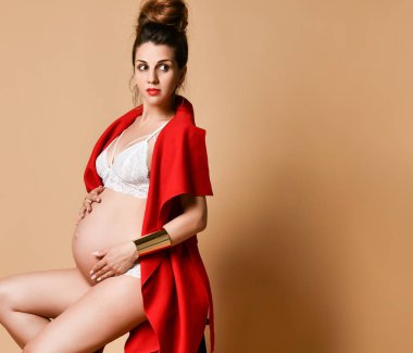 Küpeli esmer anne, bilezik, beyaz iç çamaşırı ve kırmızı şal. Sandalyeye yan oturup, bej arka planda poz vermek. Yakın plan.