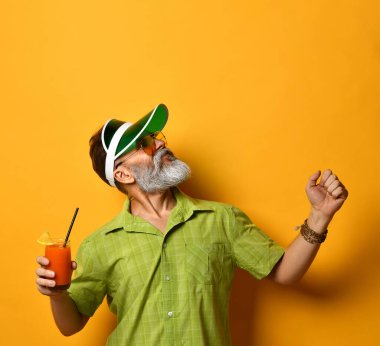 Yeşil güneşlik, gömlek ve güneş gözlüğü takan sakallı bir erkek. Bir bardak sıkılmış meyve suyu, sıkılmış yumruk, turuncu arka planda poz.