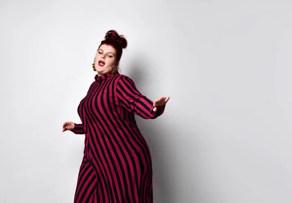 Obese redhead lady in striped dress and earrings. She reaching her hand to you while posing sideways isolated on white background — Stockfoto
