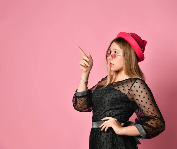 Ragazza adolescente in occhiali da sole, abito nero e cappello rosso. Indica qualcosa con l'indice, posando su sfondo rosa. Da vicino. — Foto Stock