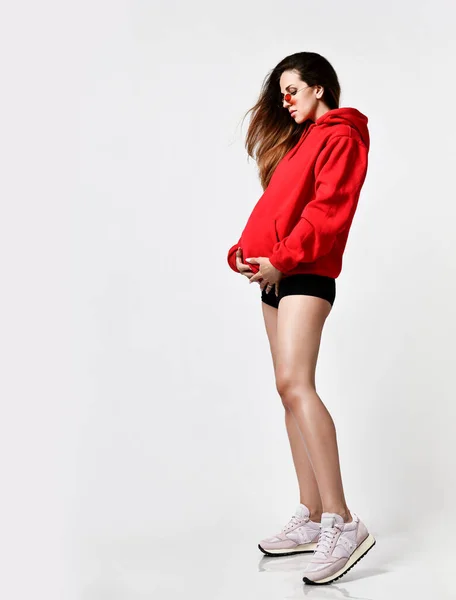 Senhora grávida em calções pretos, capuz vermelho e óculos de sol, tênis rosa. Ela está vestindo um capuz, isolado em branco. Comprimento total — Fotografia de Stock
