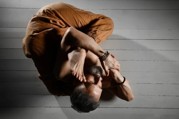 Hombre yogui envejecido con el torso desnudo, en pantalones y pulseras marrones está realizando su flexibilidad. Acostado en un suelo de madera blanca — Foto de Stock