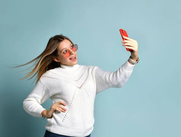 Adolescente en gafas de sol naranjas, reloj, pulsera y suéter. Ella tomando selfie, sorprendida, posando sobre fondo azul. De cerca. — Foto de Stock