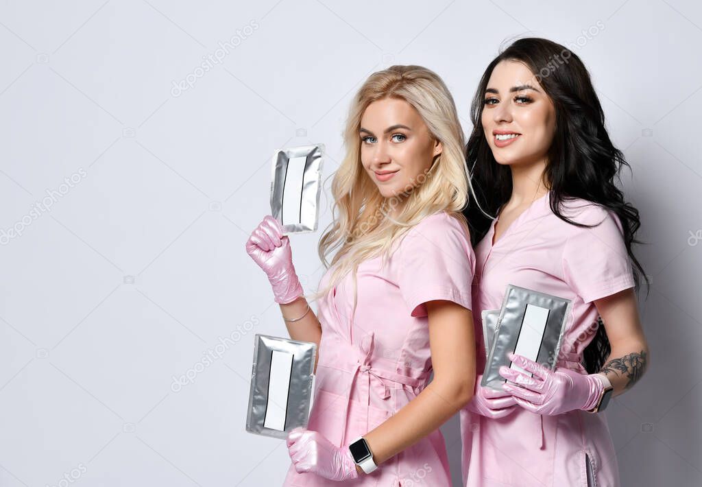Two girls in pink uniform posing isolated on white. Holding jar and bottle, cosmetic mockups for an advertising products. Close up