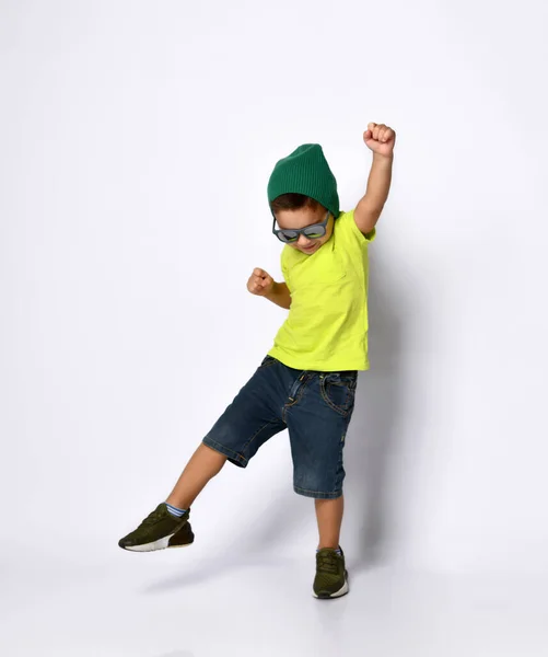 Маленький хлопчик в сонцезахисних окулярах, зелений капелюх, жовта футболка, джинсові шорти, хокейні кросівки. Піднята нога з'являється язиком. Ізольовані на білому — стокове фото