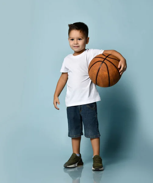 Beyaz tişörtlü, kot şortlu ve haki ayakkabılı küçük esmer çocuk. Basketbol topunu tutuyor, gülümsüyor, mavi arka planda poz veriyor. — Stok fotoğraf