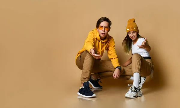 Young teens boy and girl in comfortable clothing, hats, sneakers and sunglasses sitting and pointing at camera — Φωτογραφία Αρχείου