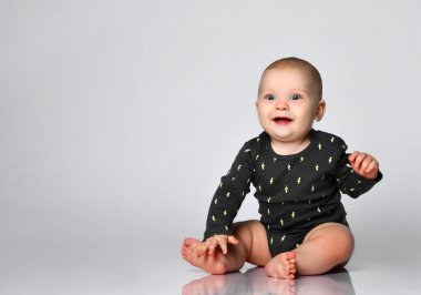 Çıplak ayaklı, siyah giysili küçük bebek. Beyaz stüdyo arka planında izole bir şekilde yerde otururken gülümsüyor. Kapat.