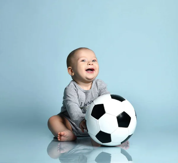 Ingefärsbarn i grå kroppsdräkt, barfota. Han håller fotboll, sitter på golvet mot blå bakgrund. Närbild, kopiera utrymme — Stockfoto