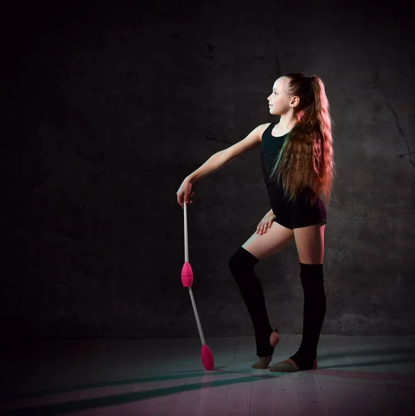 Young smiling girl gymnast in black sport body and uppers standing and holding two pink gymnastic maces in hand