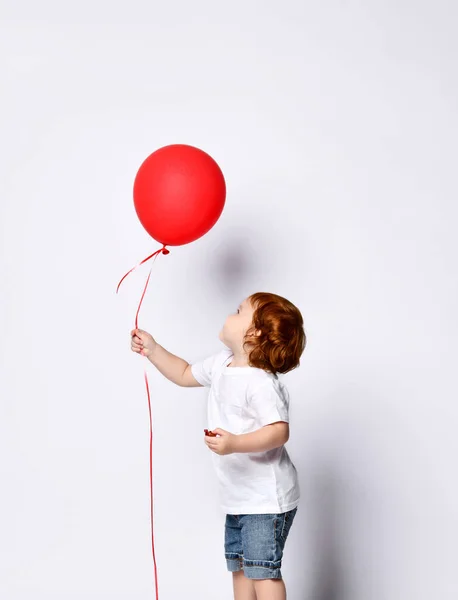Мальчик или девочка в белой футболке, туфлях, джинсовых шортах. Ребенок улыбается, держа красный воздушный шар, позируя боком изолированный на белом — стоковое фото