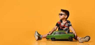 Güneş gözlüklü, kamuflaj tişörtlü, spor ayakkabılı bir çocuk. Yerde oturmuş, elinde yeşil bir eski arabanın turuncu arka planda duruşu.