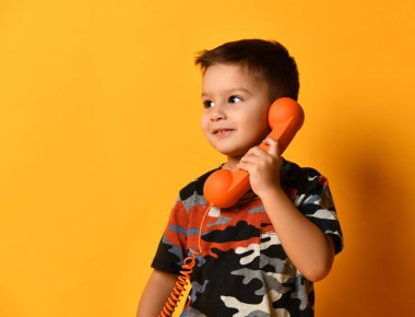 Kamuflaj tişörtlü çocuk elinde telli bir cep telefonu tutuyor, yana bakıyor ve gülümsüyor, turuncu stüdyo arka planına karşı poz veriyor.