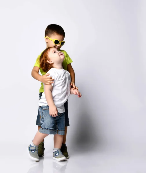 Маленький хлопчик в сонцезахисних окулярах обіймає свого молодшого брата або сестру. Вони посміхаються, одягнені в повсякденний одяг, позує ізольовано на білому — стокове фото
